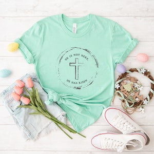Op de afbeelding: Een mintgroen t-shirt met de tekst "HE IS NOT HERE. HE HAS RISEN" gedrukt in een cirkelvormig ontwerp met een kruis. Het shirt is gestyled met een spijkerbroek, roze tulpen, paaseieren en witte sneakers.