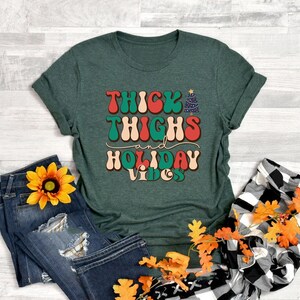 Puede incluir: Una camiseta verde con el texto "Thick Thighs and Holiday Vibes" en una fuente retro con un gr&aacute;fico de &aacute;rbol de Navidad.