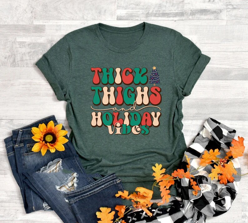 Puede incluir: Una camiseta verde con el texto "Thick Thighs and Holiday Vibes" en una fuente retro con un gr&aacute;fico de &aacute;rbol de Navidad.