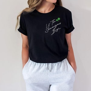Peut inclure: Un t-shirt noir avec un texte blanc qui dit "Let the shenanigans begin" avec deux tr&egrave;fles verts. Le mod&egrave;le porte le t-shirt avec un pantalon de surv&ecirc;tement gris.