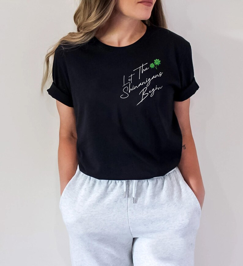 Peut inclure: Un t-shirt noir avec un texte blanc qui dit "Let the shenanigans begin" avec deux tr&egrave;fles verts. Le mod&egrave;le porte le t-shirt avec un pantalon de surv&ecirc;tement gris.