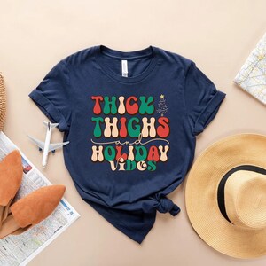 Puede incluir: Camiseta azul marino con un gr&aacute;fico de estilo retro que dice "Thick Thighs and Holiday Vibes". El gr&aacute;fico presenta colores rojo, verde y blanco.