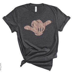 May include: A dark grey t-shirt with a rose gold glitter hand making the shaka sign.