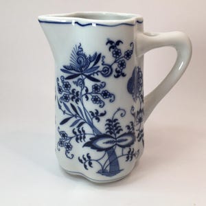 May include: A white porcelain creamer pitcher featuring a blue onion pattern with intricate floral and botanical motifs. The pitcher has a scalloped rim, a curved handle, and a smooth glazed finish with blue painted details along the edges.