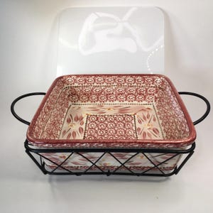 May include: Square ceramic baking dish with a floral pattern in red and cream, set in a black metal wire frame. The dish has black handles and a white lid. A decorative kitchenware item.