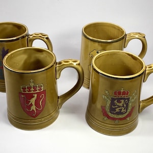 May include: Four ceramic mugs with gold handles and rims. The mugs are a muted gold color and feature various heraldic crests in red, gold, and purple. The mugs are cylindrical with a slightly flared top.
