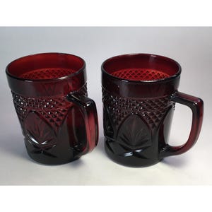 May include: Two ruby red glass mugs with a diamond and leaf pattern. Each mug has a handle and a textured interior. The mugs are made of a dark red glass and are set against a white background.