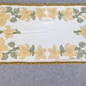 May include: White rectangular table runner with embroidered yellow lilies and green leaves. The edges are trimmed with a gold-colored, scalloped border. A decorative floral accent for a dining table.