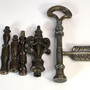 May include: An assortment of antique brass hardware components, including finials and a key-shaped arrow. The finials vary in shape and size, with threaded bases. The arrow key features a loop handle and detailed feather fletching. The items are displayed on a white surface.