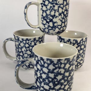 May include: Four ceramic mugs with a blue and white speckled pattern. Each mug has a rounded body, a large handle, and a cream-colored interior. The mugs are stacked and arranged against a white background.