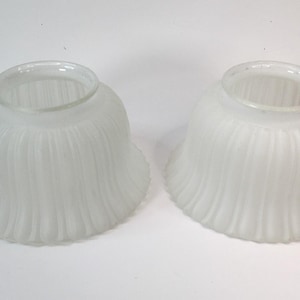 May include: A pair of bell-shaped lamp shades made of frosted white glass. Each shade features a vertical ribbed texture along the exterior surface, a wide flared opening at the bottom, and a smooth, clear glass circular fitter at the top.