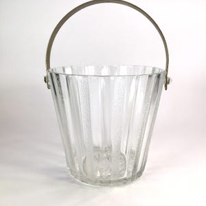 May include: A clear glass ice bucket with vertical textured stripes and a metal handle. The bucket has a flared shape and a decorative pattern at the base. The handle is arched and attached to the bucket with small metal brackets.