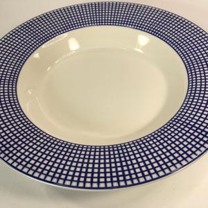 May include: A white ceramic soup bowl with a blue checkered border. The bowl has a wide rim with a repeating grid pattern in navy blue. The center of the bowl is plain white, creating a contrast with the patterned edge.