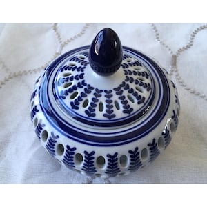 May include: A white and blue ceramic jar with a lid. The jar has a round shape with a series of oval-shaped openings around the sides. The lid is decorated with a blue floral pattern and a dark blue knob. The jar is likely used for decorative purposes.