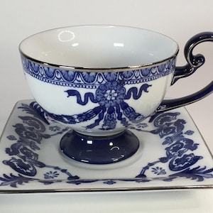 May include: White teacup and saucer set with a blue floral design and silver trim. The teacup has a curved handle and sits on a square saucer. The design includes ribbons and floral patterns.