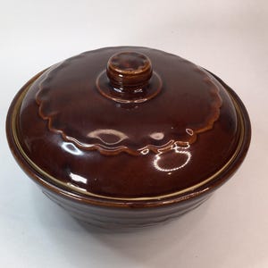 May include: A brown ceramic casserole dish with a lid. The dish has a round shape with a scalloped edge on the lid and a small knob handle. The exterior is a glossy, dark brown color.