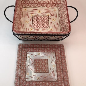 May include: A square, ceramic baking dish with a floral pattern in red and cream, set in a black metal wire frame. A matching square glass lid with a similar design is next to the dish. The dish has two curved handles.