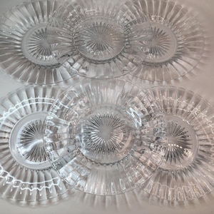May include: A set of five clear glass plates arranged in two rows. Each plate features a circular center with a radiating sunburst pattern, surrounded by a wide, smooth rim with vertical fluted ridges extending to the outer edge.
