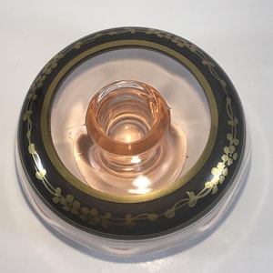 May include: A round, pale pink glass candle holder featuring a wide, dark band around the top rim. The dark band is decorated with a delicate, repeating gold floral vine pattern. The center of the holder has a circular opening for a candle.