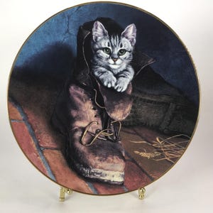 May include: Decorative plate featuring a gray tabby kitten nestled inside a worn brown boot. The plate has a gold rim and a detailed illustration of the cat and boot on a brick and blue background. A charming cat-themed collectible.