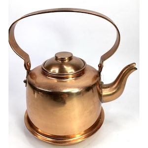 May include: A copper-colored kettle with a large, arched handle and a spout. The kettle has a round base and a lid with a small knob. The handle is attached to the body of the kettle with rivets. The kettle is made of a shiny metal.