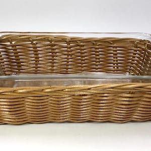 May include: A rectangular glass baking dish inside a woven wicker basket. The basket is a light brown color and has a handle on each end. The glass dish is clear and has rounded corners.