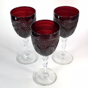 May include: Three ruby red wine glasses with intricate diamond and leaf patterns. The glasses have clear stems and bases, creating a striking contrast with the deep red bowls. The elegant design suggests a vintage or antique style, perfect for special occasions.