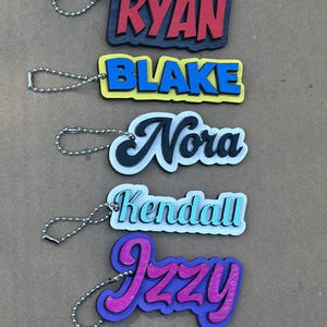 May include: A collection of personalized name keychains. Each keychain features a different name in a colorful, stylized font. The names include "Lilah", "Ryan", "Blake", "Nora", "Kendall", "Izzy", and "Liv". Each keychain has a silver ball chain.