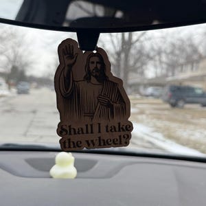 May include: Wooden car air freshener featuring an image of Jesus with his hand raised in a blessing. The text "Shall I take the wheel?" is below the image. A small, yellow duck-shaped item is visible below the air freshener.