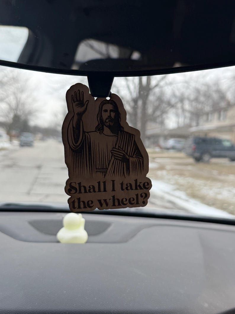 May include: Wooden car air freshener featuring an image of Jesus with his hand raised in a blessing. The text "Shall I take the wheel?" is below the image. A small, yellow duck-shaped item is visible below the air freshener.