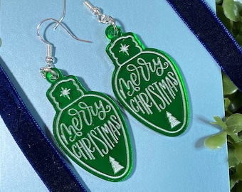 May include: Green Christmas light-shaped earrings with the words "Merry Christmas" in white script. The earrings have silver hooks and a small Christmas tree design. The earrings are displayed on a light blue card, with a green leafy background and blue ribbon.