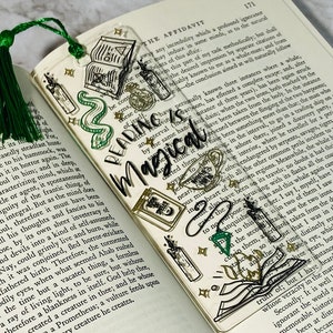 May include: A clear bookmark with a green, black, and gold design. The bookmark features a snake, a book, a teacup, a candle, and a quill. The text "Reading is Magical" is written in a white, cursive font.