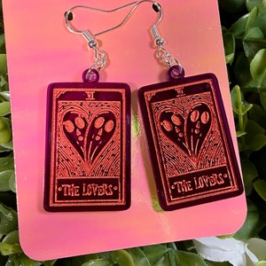 May include: A pair of dangle earrings featuring a tarot card design. The earrings are rectangular with a dark purple background and a red design of a heart with two faces. The text "VI" and "THE LOVERS" are visible.