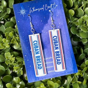 May include: A pair of silver dangle earrings with white rectangular charms that say "CUBAN BREAD" in blue and red lettering. The earrings are displayed on a blue and white background with the text "Whimsical Craft Co."