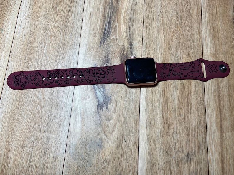 May include: A maroon smartwatch with a gold-colored case and a matching maroon band. The band is decorated with black medical-themed illustrations. The watch is displayed on a wooden surface.