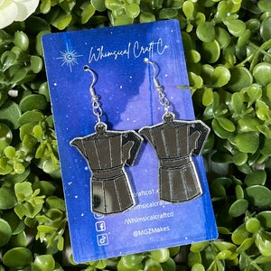 May include: Silver-colored coffee pot earrings with a reflective surface, hanging from silver hooks. The earrings are displayed on a blue card with the text "Whimsical Craft Co" and social media handles. The background is a bed of green foliage.
