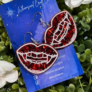 May include: Red glitter vampire teeth earrings with silver hooks. The earrings are displayed on a blue background with the text "Whimsical Craft Co."
