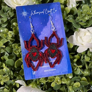 May include: A pair of red and black spider earrings with a heart on the spider's body. The earrings are on a white card with the text "Whimsical Craft Co." and "@MGZMakes".