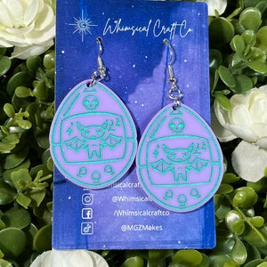 May include: A pair of lavender earrings with a teal bat design inside a retro-style egg shape. The earrings have silver hooks and are displayed on a blue card with the text "Whimsical Craft Co."