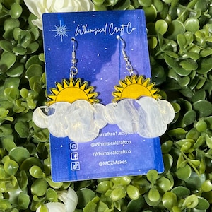 May include: A pair of earrings featuring a white cloud with a yellow sun behind it. The earrings are on a card with the text "Whimsical Craft Co" and social media handles.