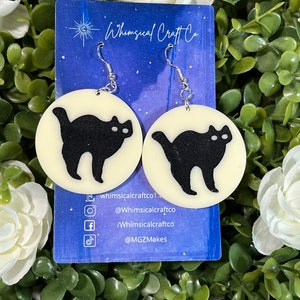 May include: A pair of earrings featuring a black cat silhouette on a white circular background. The earrings are on a card with the text "Whimsical Craft Co." and social media handles.