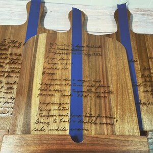 May include: Three wooden cutting boards with handwritten recipes for banana bread cake, roast, and a recipe for a dessert. The boards are engraved with the recipes and have blue tape on the back.