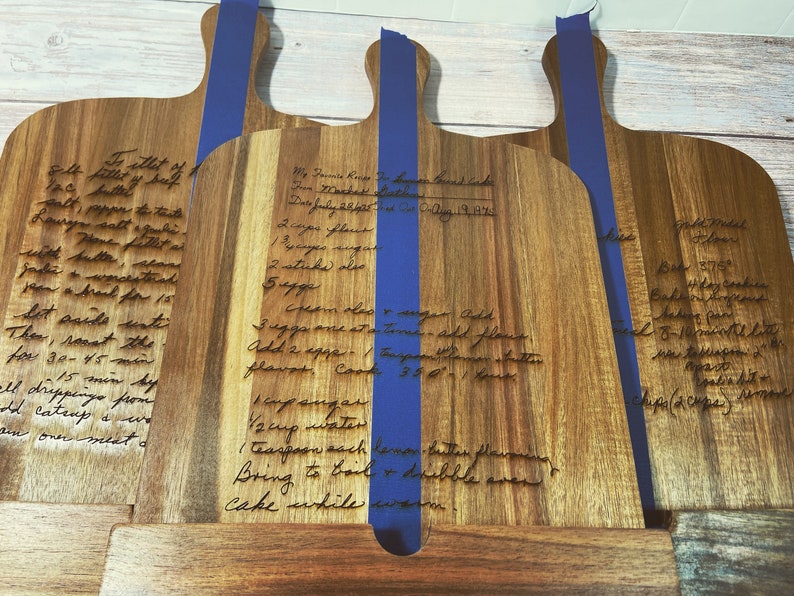 May include: Three wooden cutting boards with handwritten recipes for banana bread cake, roast, and a recipe for a dessert. The boards are engraved with the recipes and have blue tape on the back.