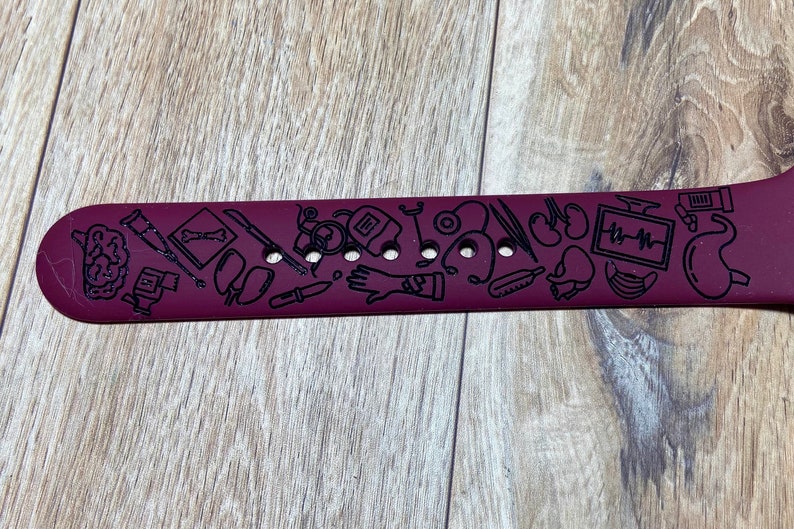 May include: A maroon watch band with black etched medical-themed illustrations. The band features images of a brain, bones, medical tools, and organs. The band has multiple holes for adjustment.