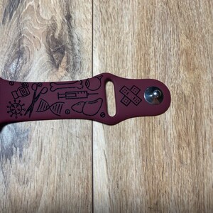 May include: A maroon watch band with black medical-themed illustrations. The band features a variety of medical symbols, including a virus, scissors, a DNA strand, a syringe, and bandages. The band is made of a flexible material.