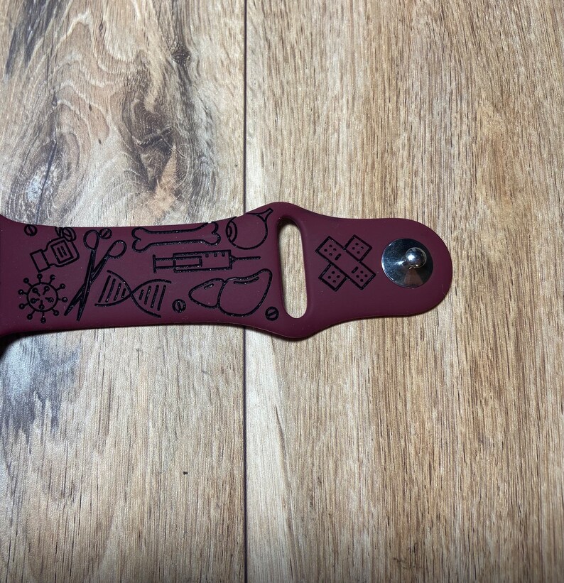 May include: A maroon watch band with black medical-themed illustrations. The band features a variety of medical symbols, including a virus, scissors, a DNA strand, a syringe, and bandages. The band is made of a flexible material.