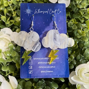 May include: White and gold marbled cloud earrings with gold lightning bolt accents. The earrings are hanging from silver hooks. The earrings are displayed on a blue background with the text "Whimsical Craft Co." and social media handles.