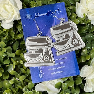 May include: A pair of silver-colored earrings shaped like a stand mixer. The earrings are on a blue and white background with the text "Whimsical Craft Co." and social media handles.