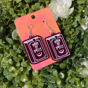 May include: A pair of dangle earrings featuring a burgundy book design with the words "BOOK OF SPELLS" in white lettering. The earrings have a crescent moon symbol and silver-colored fish hook ear wires. The earrings are displayed on a pink and orange background.
