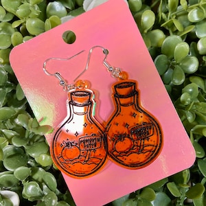 May include: A pair of orange pumpkin spice potion bottle earrings. The earrings are made of clear acrylic with a black outline and a pumpkin spice sign.